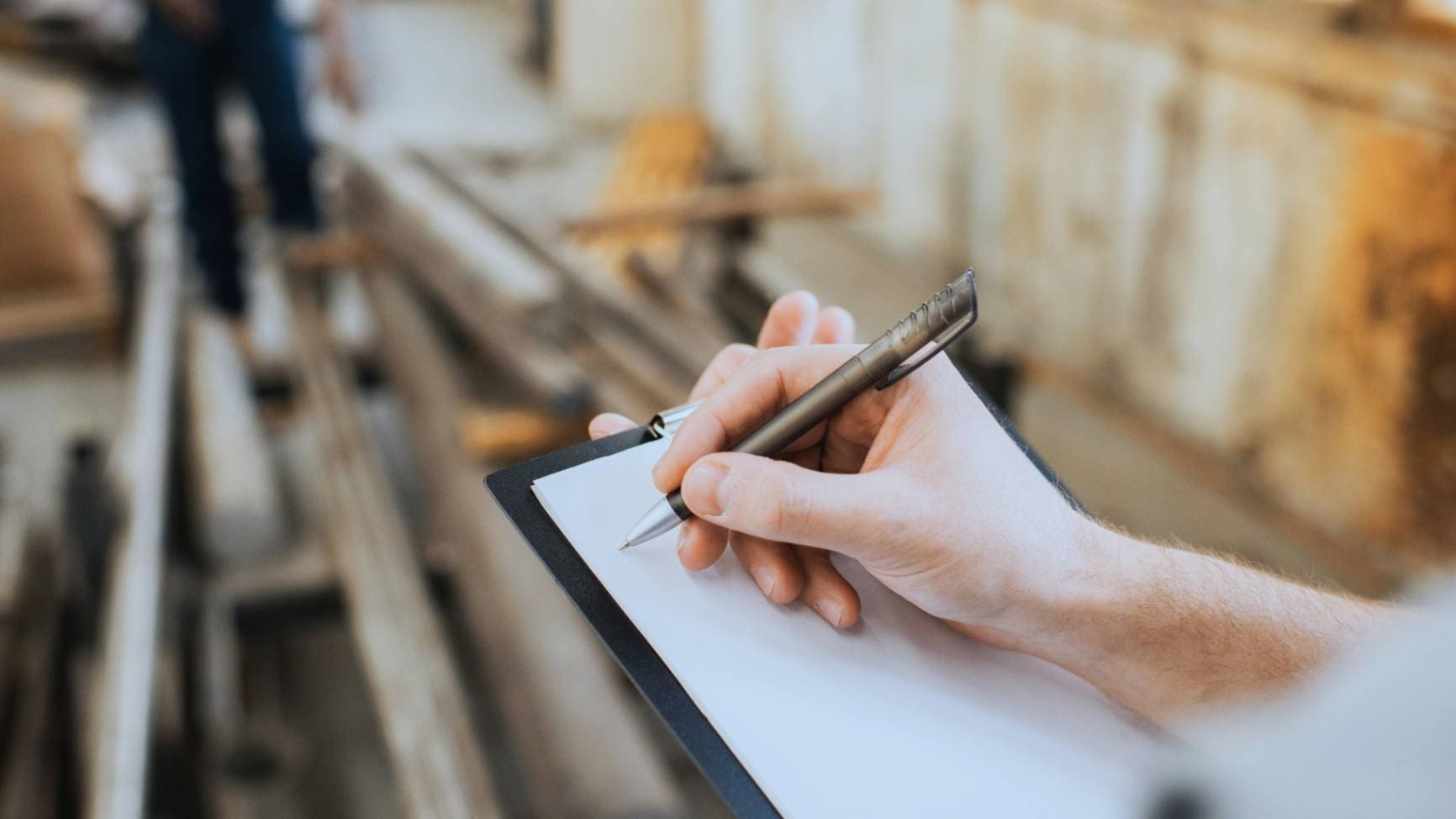 Close-up of a person's hand holding a pen over a clipboard in an industrial setting, evaluating materials.