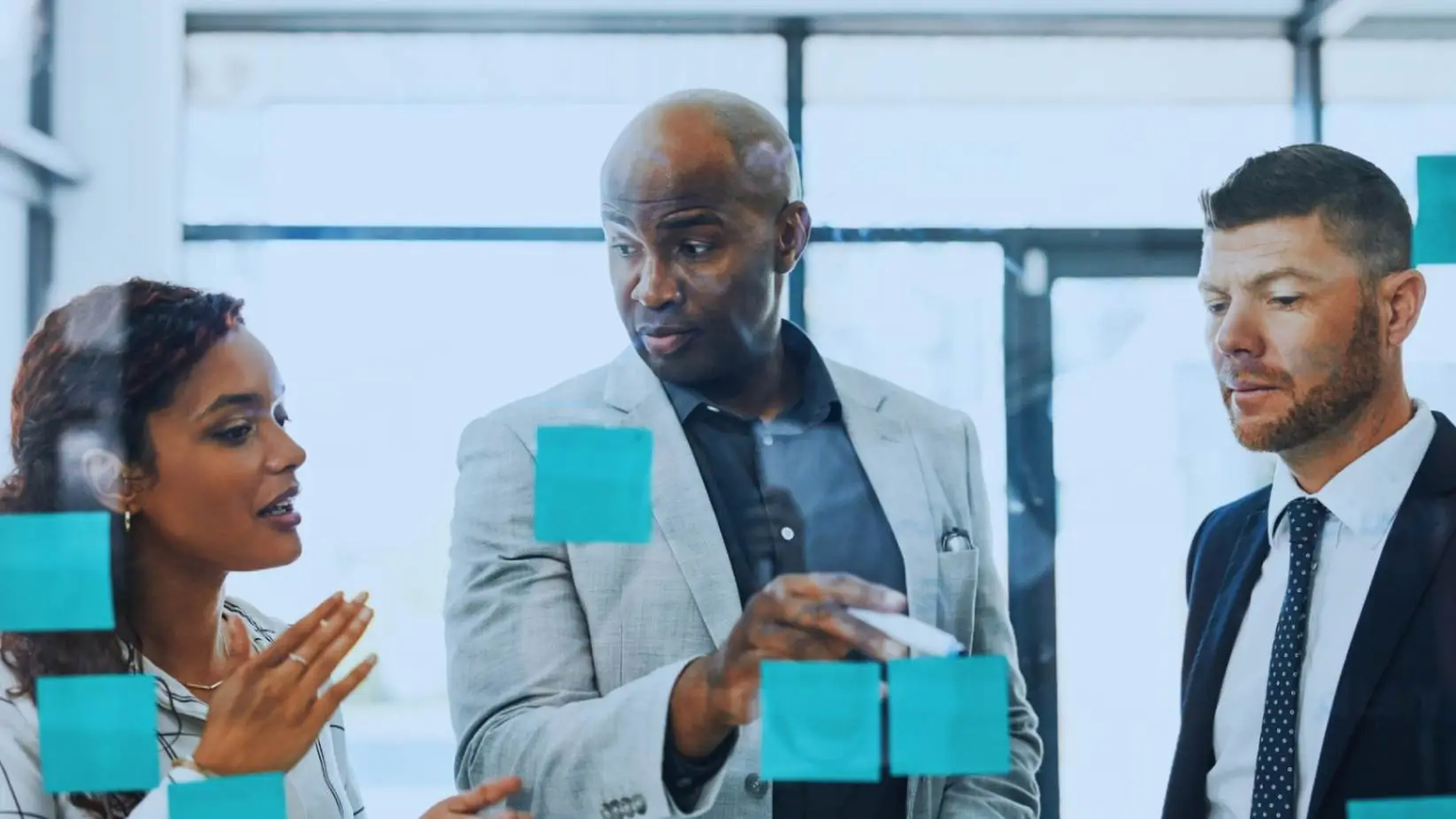 "Diverse corporate team discussing project details with sticky notes on a glass board, including African American and Caucasian business professionals in a light-filled office."