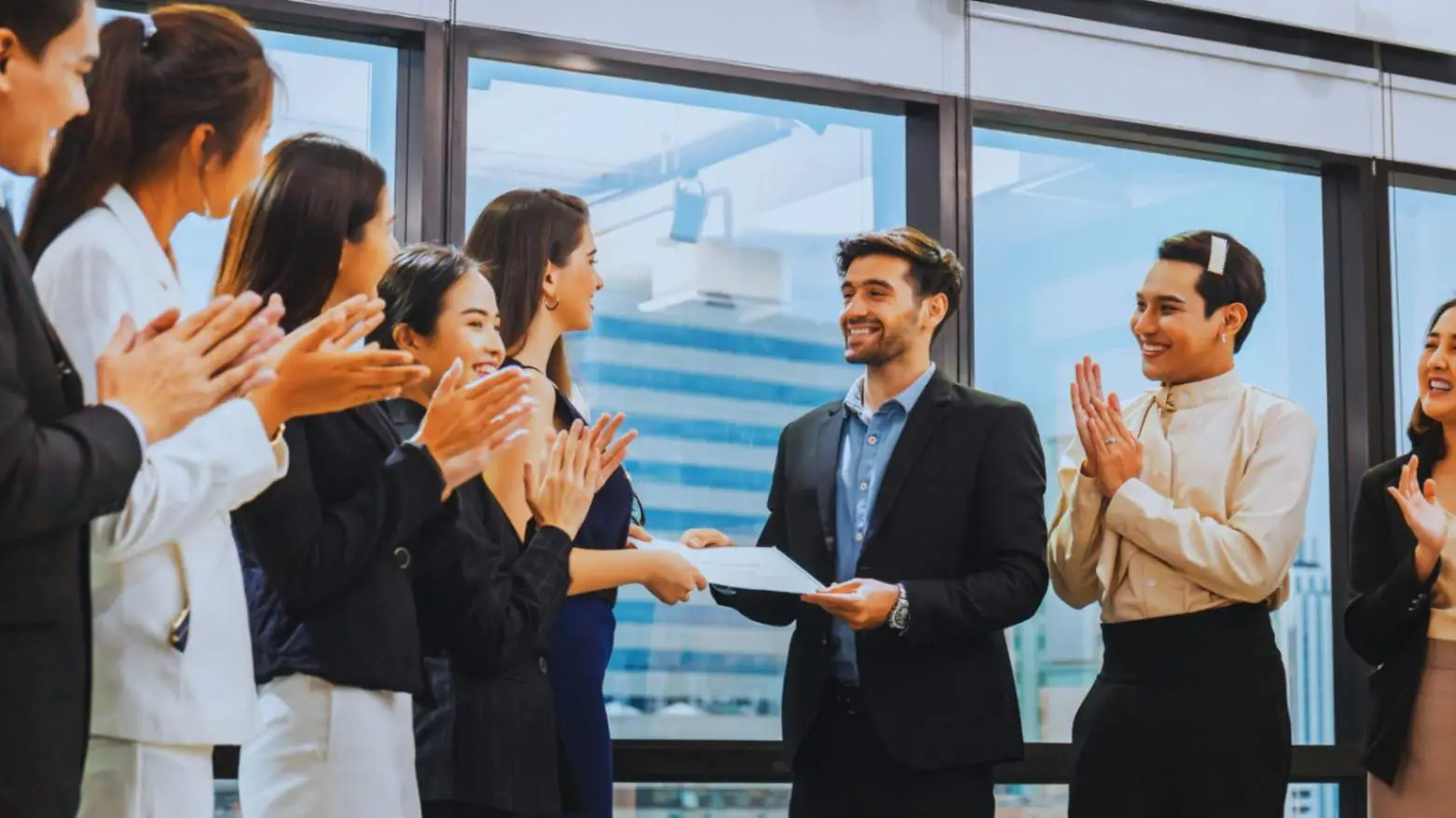 "Group of Asian and Hispanic colleagues applauding a young Caucasian man receiving an award in a modern office, celebrating teamwork and achievement."